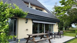 Une maison dotée d’un patio comprenant une table et des chaises en bois pour pique-niquer, de grandes fenêtres et une vue sur la verdure environnante.