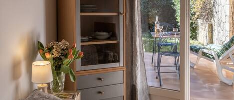 Une chambre avec un lit, une table de chevet avec une lampe et un vase de fleurs, une armoire contenant de la vaisselle, et une vue sur une terrasse extérieure avec une table et des chaises.