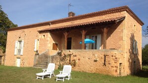 Un edificio rústico de piedra con techo de tejas, dos sillones blancos y una sombrilla azul en el porche.