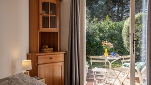 A bedroom with a bed, a wooden cabinet, a nightstand with a lamp, and a view of an outdoor patio with a table, chairs, and a garden. - Charming studio in villa in Saint Paul de Vence with terrace and garden (Saint-Paul-De-Vence)