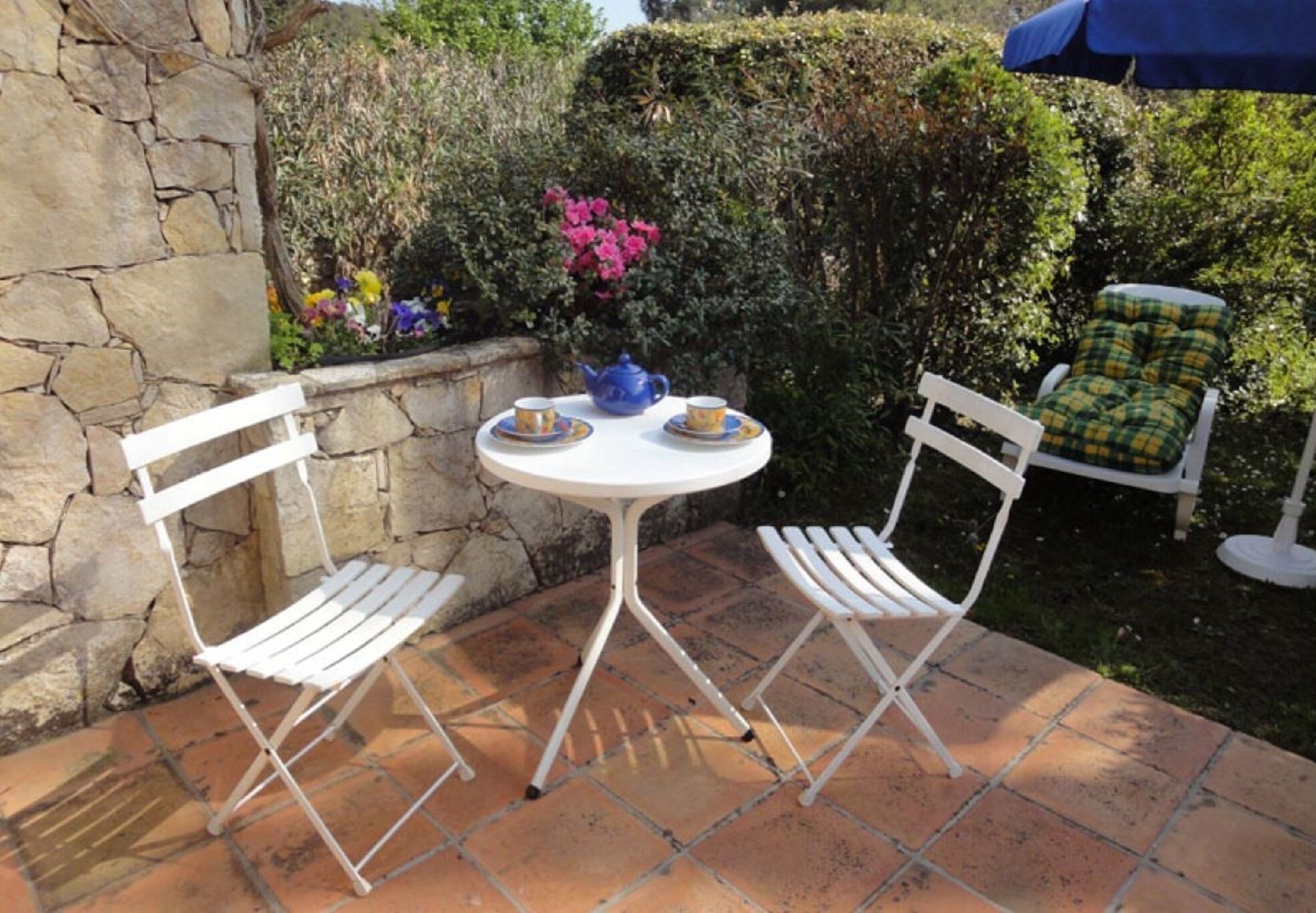 Une terrasse avec une table blanche et deux chaises blanches, une théière bleue et un vase rempli de fleurs.