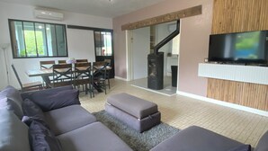 Een moderne woonkamer met een modulaire bank, een eethoek met stoelen en een tafel, een tv gemonteerd aan een houten paneel, en een open haard.