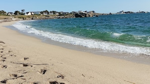 En sandstrand med bløde bølger, en klippefyldt kystlinje og huse i baggrunden.