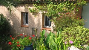 A rustic building with a tiled roof, two windows with shutters, and a garden with various plants and flowers. - In villa, charming studio 2 pers with terrace in St Paul de Vence (Saint-Paul-De-Vence)