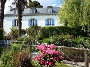Un bâtiment blanc avec des volets bleus et un jardin où poussent des fleurs roses.