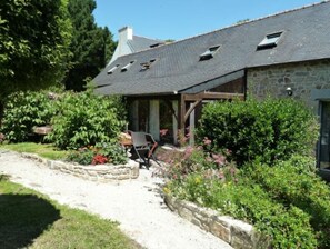 Une maison en pierre avec un toit en pente, un porche abrité et un jardin où poussent différentes plantes et fleurs.
