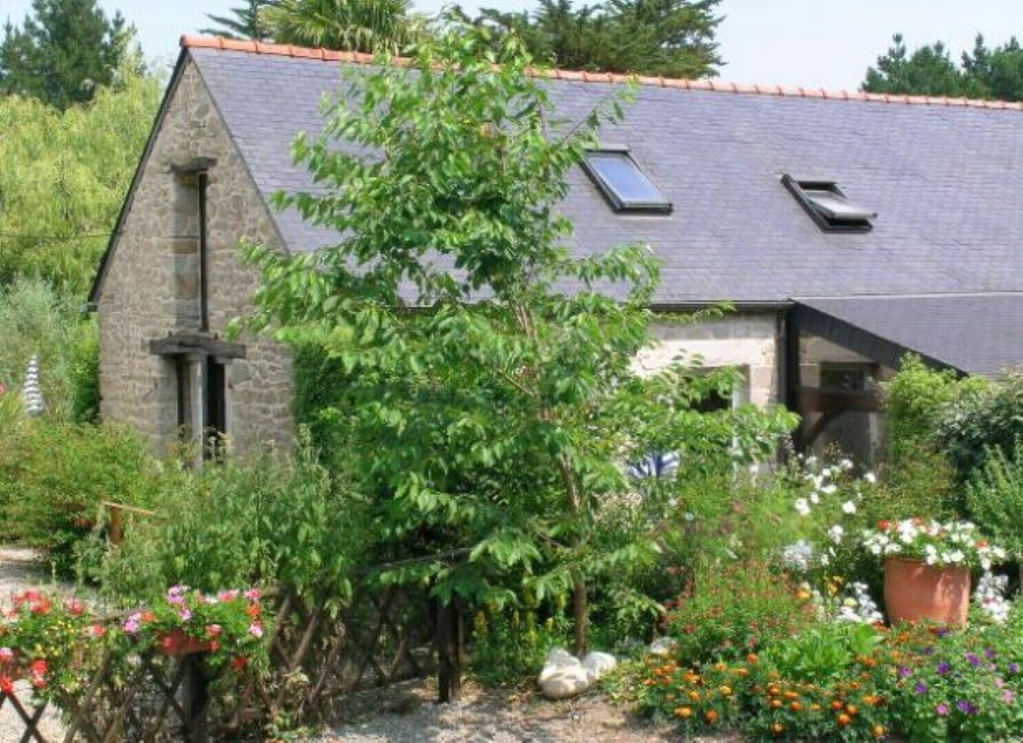 Une maison en pierre avec un toit en pente, une cheminée et un jardin où poussent différentes plantes et fleurs.