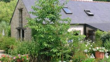 Une maison en pierre avec un toit en pente, une cheminée et un jardin où poussent différentes plantes et fleurs.