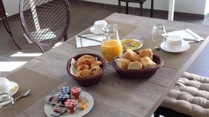Une table en bois, dans une pièce moderne, est dressée pour le petit-déjeuner avec des croissants, des pâtisseries et du jus de fruits. On aperçoit une chaise et une petite table au fond de la pièce.