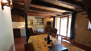 Un coin repas avec une table dressée pour deux personnes, un ordinateur portable et une bouteille de vin. On aperçoit, au fond, une cuisine avec des meubles en bois et un téléviseur.