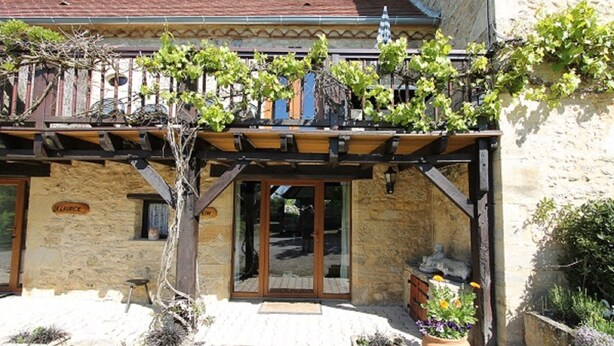 Un bâtiment en pierre surmonté d’une pergola en bois recouverte de vignes. On y trouve des plantes en pot et un petit espace de détente extérieur.