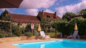 Une piscine entourée d’un jardin et d’un bâtiment dont le toit est recouvert de tuiles rouges, avec des chaises longues et des parasols à proximité.
