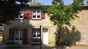 Un bâtiment à deux étages, doté de volets rouges, d’une façade en pierre et d’un petit espace extérieur aménagé avec une table et des chaises.