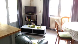 Un salon avec un canapé en cuir, une table basse sur laquelle se trouve une assiette de nourriture, un téléviseur posé sur un meuble, une bibliothèque et une table à manger avec des chaises.