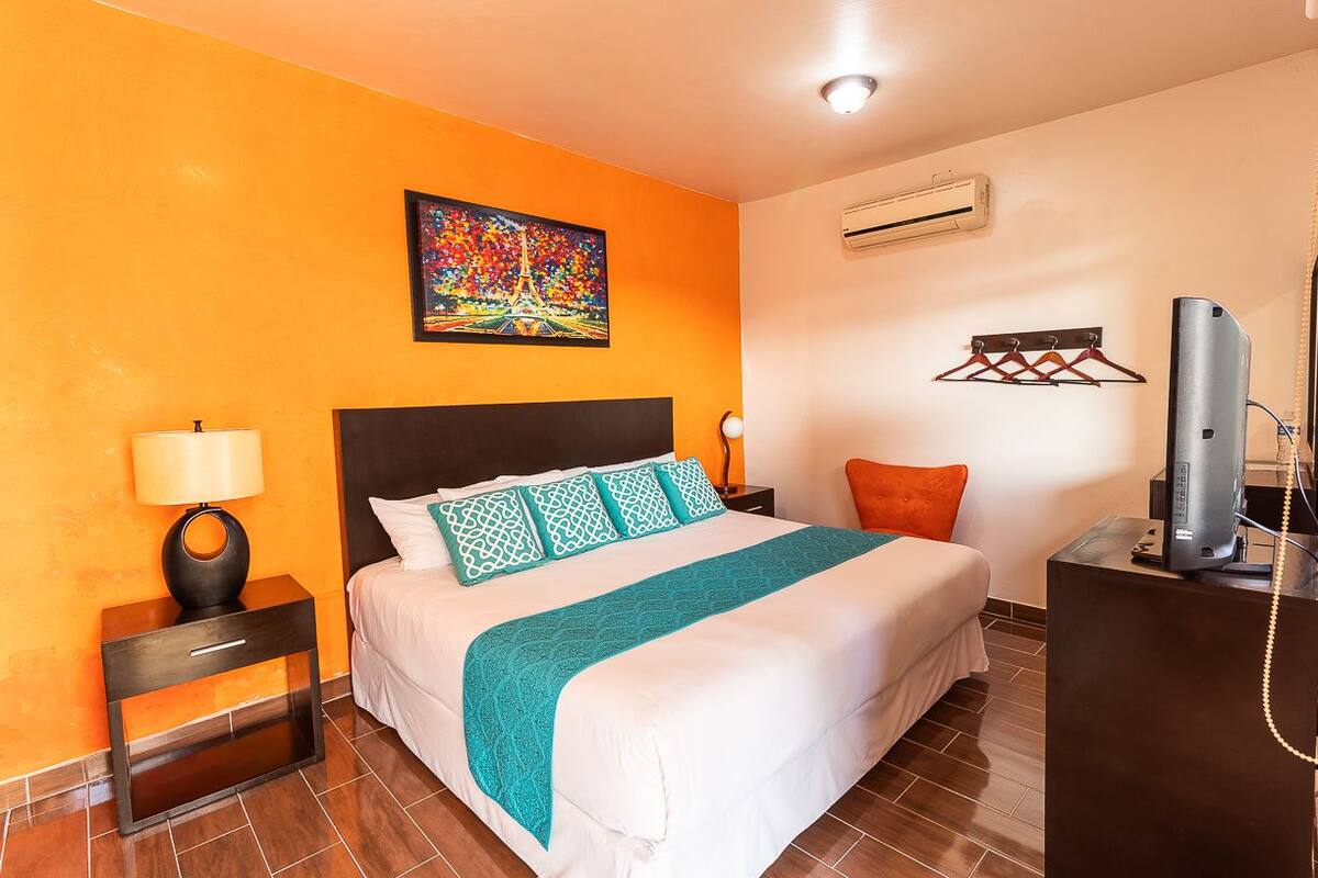 A hotel room with a bed, a nightstand with a lamp, a TV, and a painting on the wall.