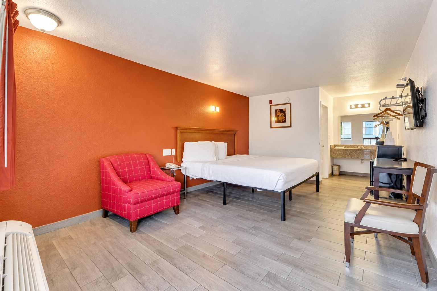 A hotel room with a bed, a red armchair, a desk with a chair, a small kitchenette, and a bathroom with a shower.