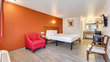A hotel room with a bed, a red armchair, a desk with a chair, a small kitchenette, and a bathroom with a shower.