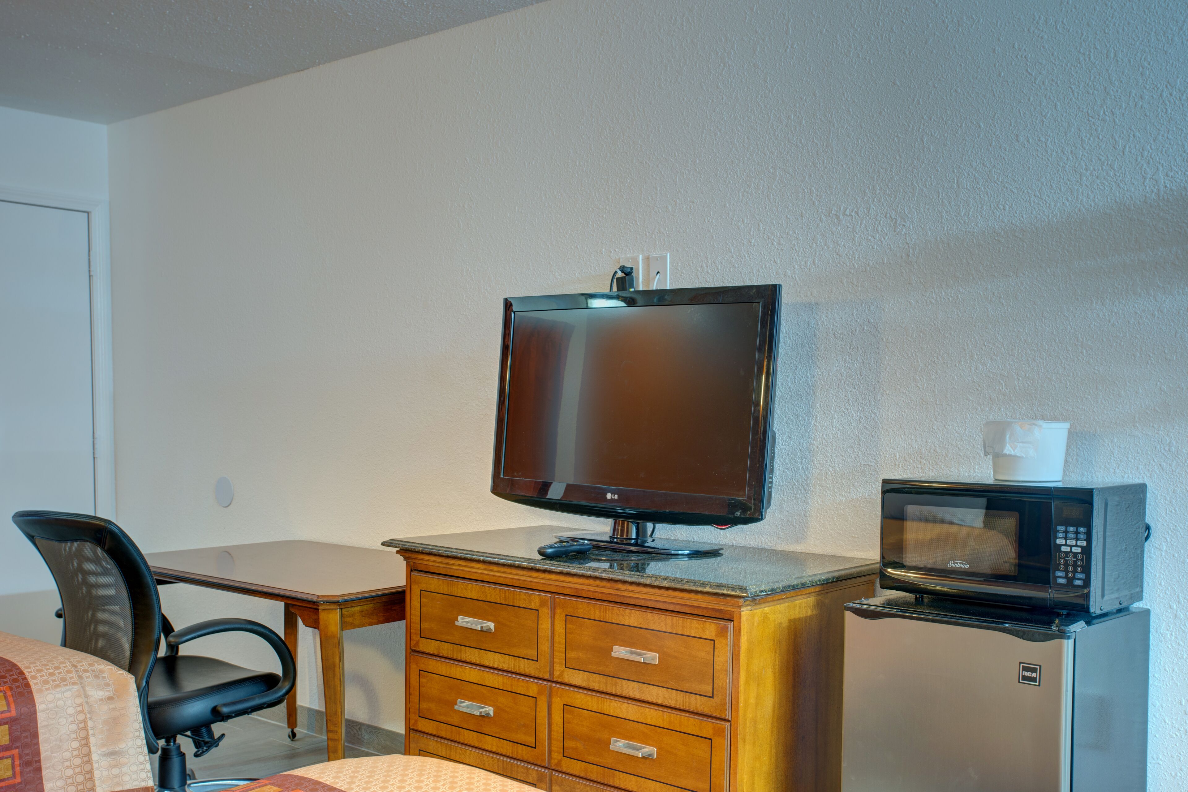 A hotel room with a desk, a chair, a television, a microwave, and a refrigerator.