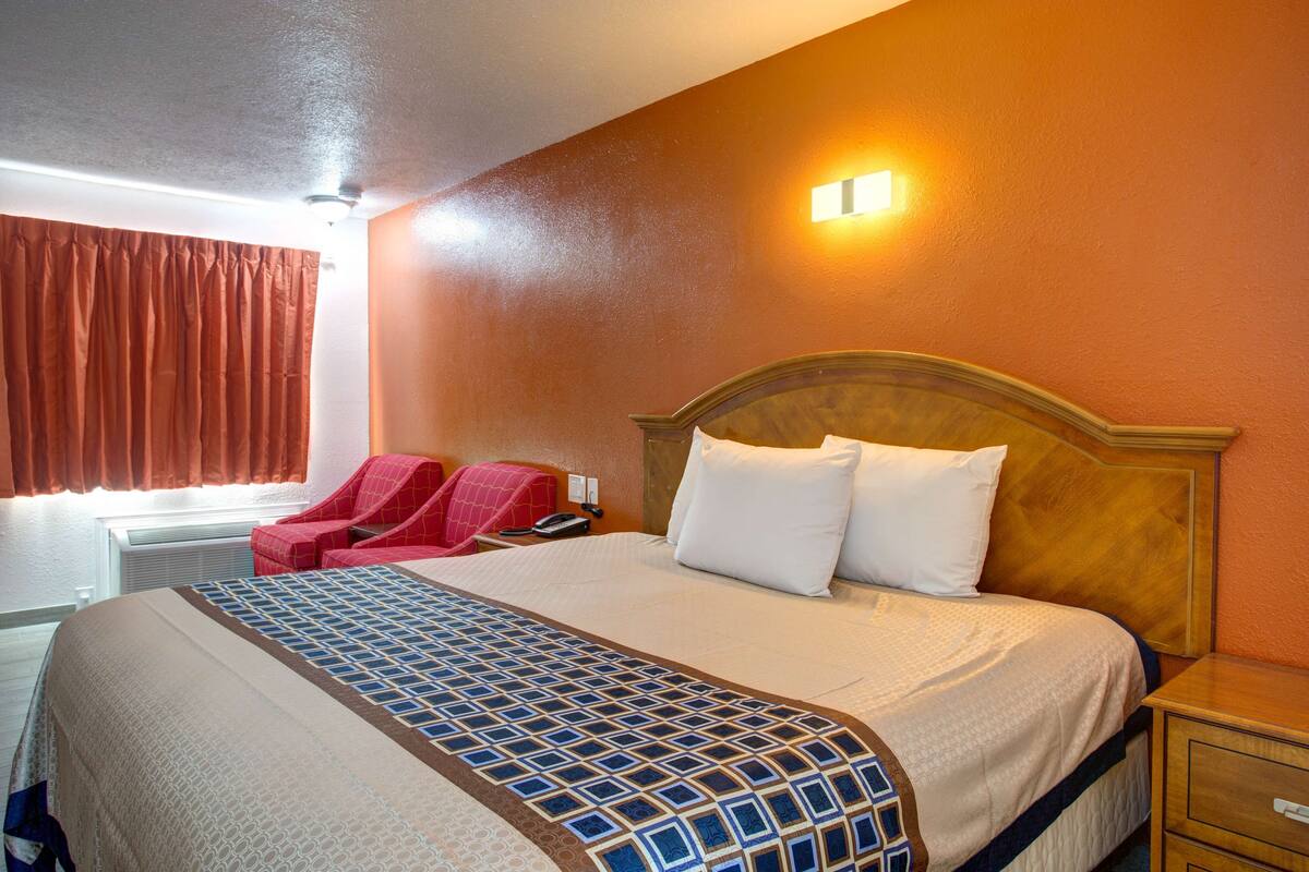 A hotel room with a large bed, a wooden headboard, a red armchair, and a nightstand with a lamp.
