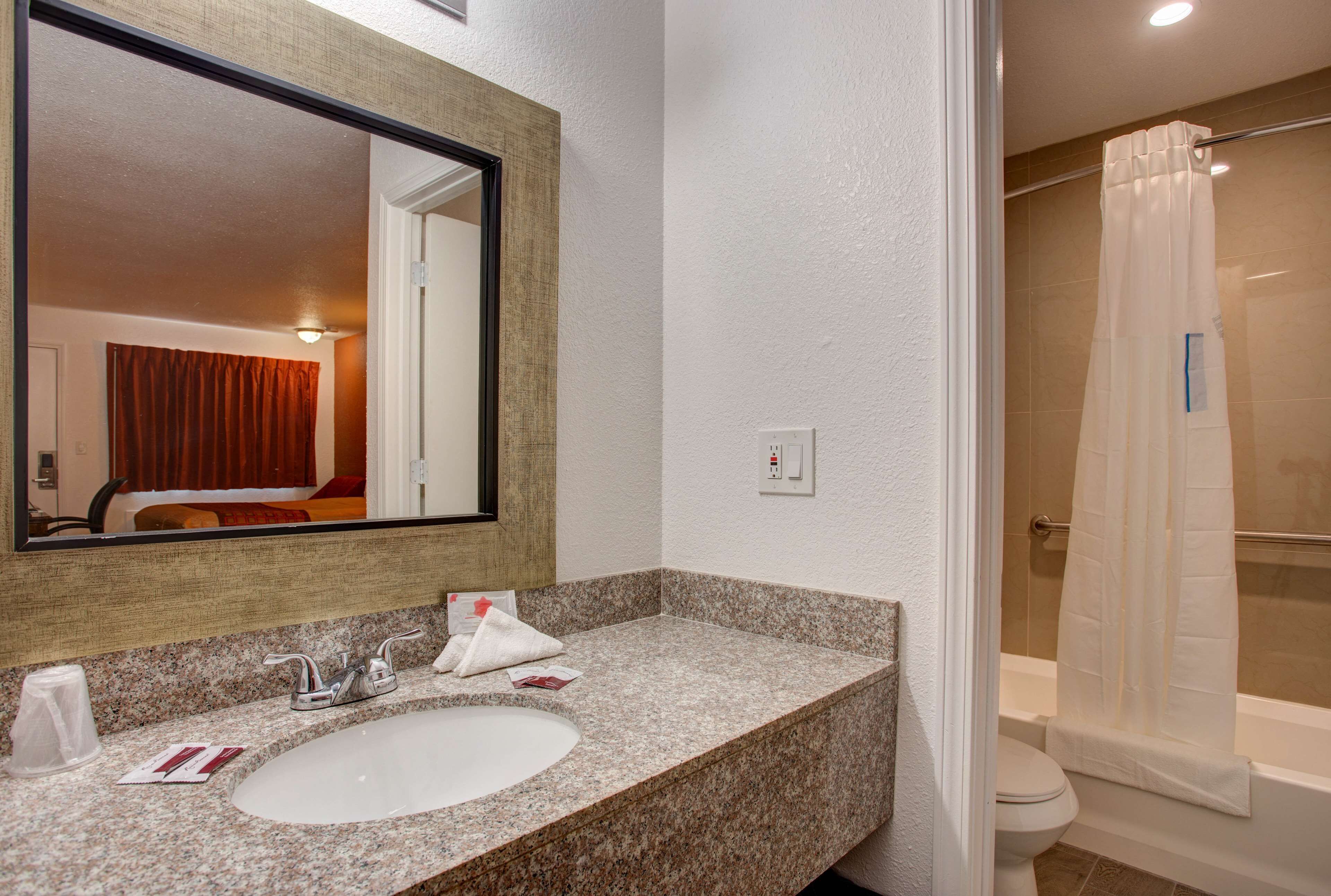 Une salle de bain d’hôtel avec un lavabo doté d’un plan de travail en granit, un grand miroir, un rideau de douche et des toilettes bien visibles.