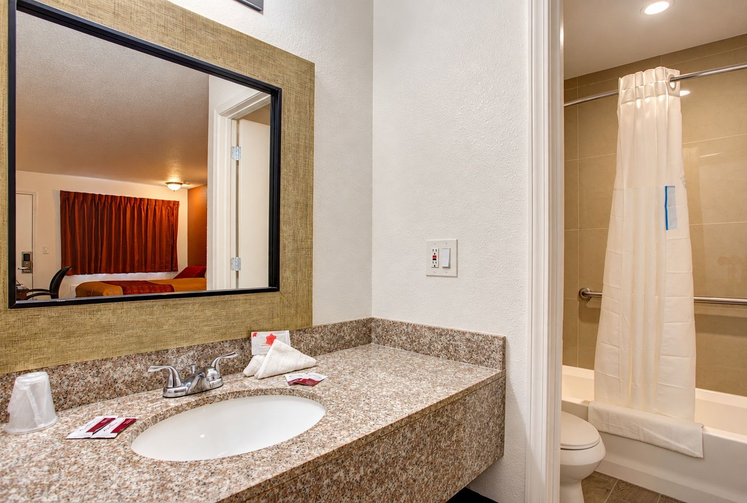 A hotel bathroom with a granite countertop sink, a large mirror, a shower with a white curtain, and a visible toilet.