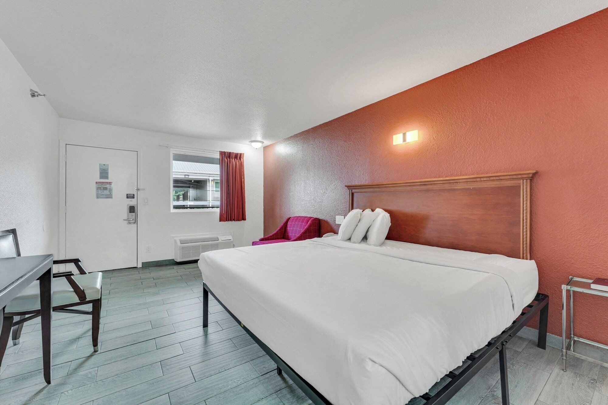 Ein Hotelzimmer mit Bett, Schreibtisch, Stuhl, einem Fenster mit Vorhängen und einer Tür mit einem Schild.