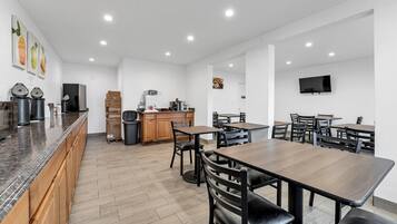 A modern dining area with wooden tables and chairs, a kitchen in the background, and a flat-screen TV mounted on the wall.