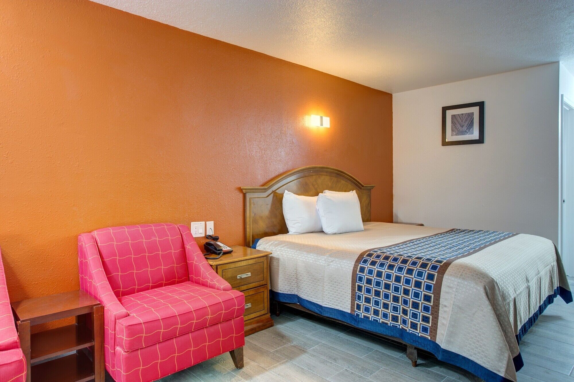 Une chambre d’hôtel avec un lit, une table de chevet en bois, un fauteuil rose et une petite table en bois.