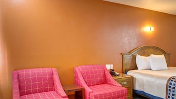 A hotel room with a bed, two pink chairs, a wooden nightstand, and a wall-mounted light.