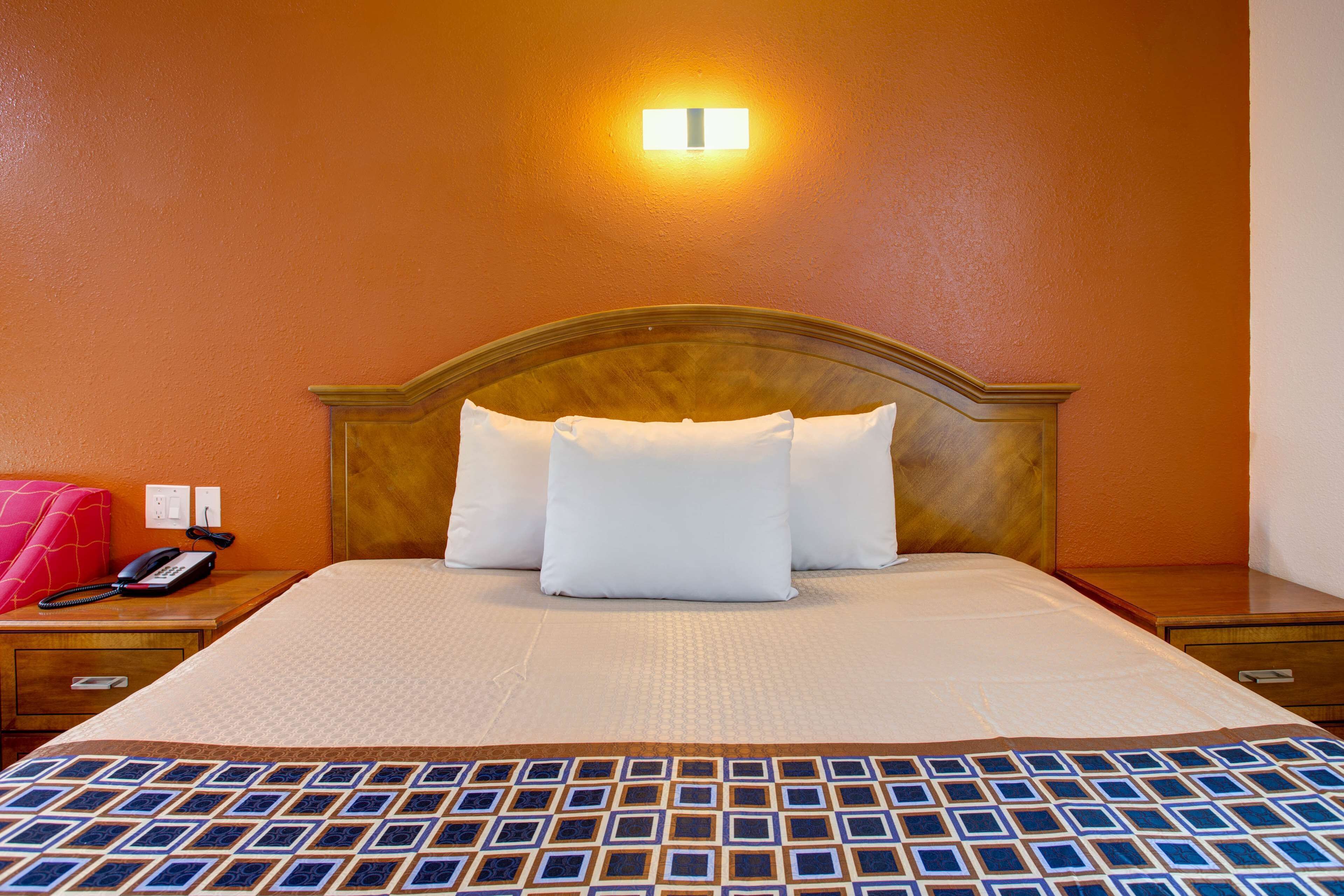 A hotel room with a wooden headboard, a bed with white linens, a patterned bedspread, a bedside table with a telephone, and a wall-mounted light fixture.