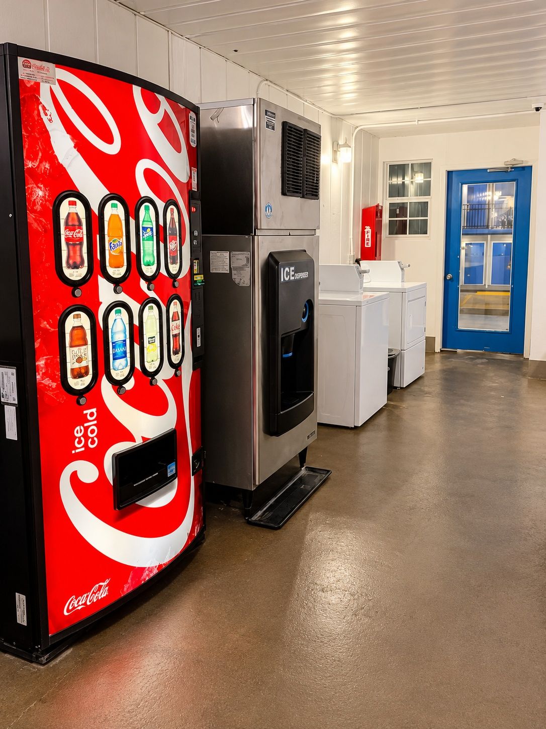 Un distributeur automatique proposant diverses boissons, un appareil à glaçons à usage commercial et une porte bleue dans un espace commercial.