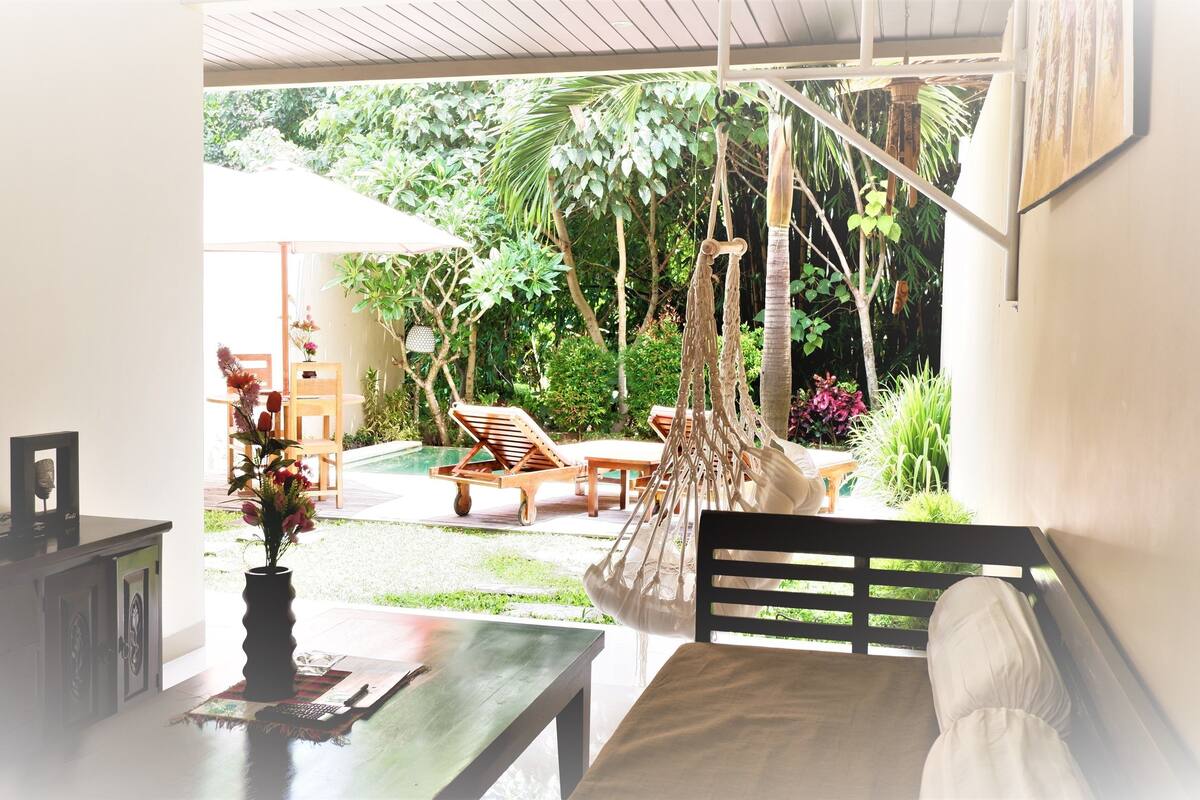 A room with a bed, a desk, and a view of an outdoor garden with a pool and lounge chairs.
