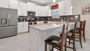 A modern kitchen with white cabinets, a central island with bar stools, stainless steel appliances, and a red window valance.