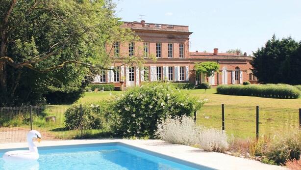 A swimming pool with a swan float, surrounded by a well-maintained lawn and a large brick building in the background. - Studio Colonel - Sleeps 4 (Castelsarrasin)