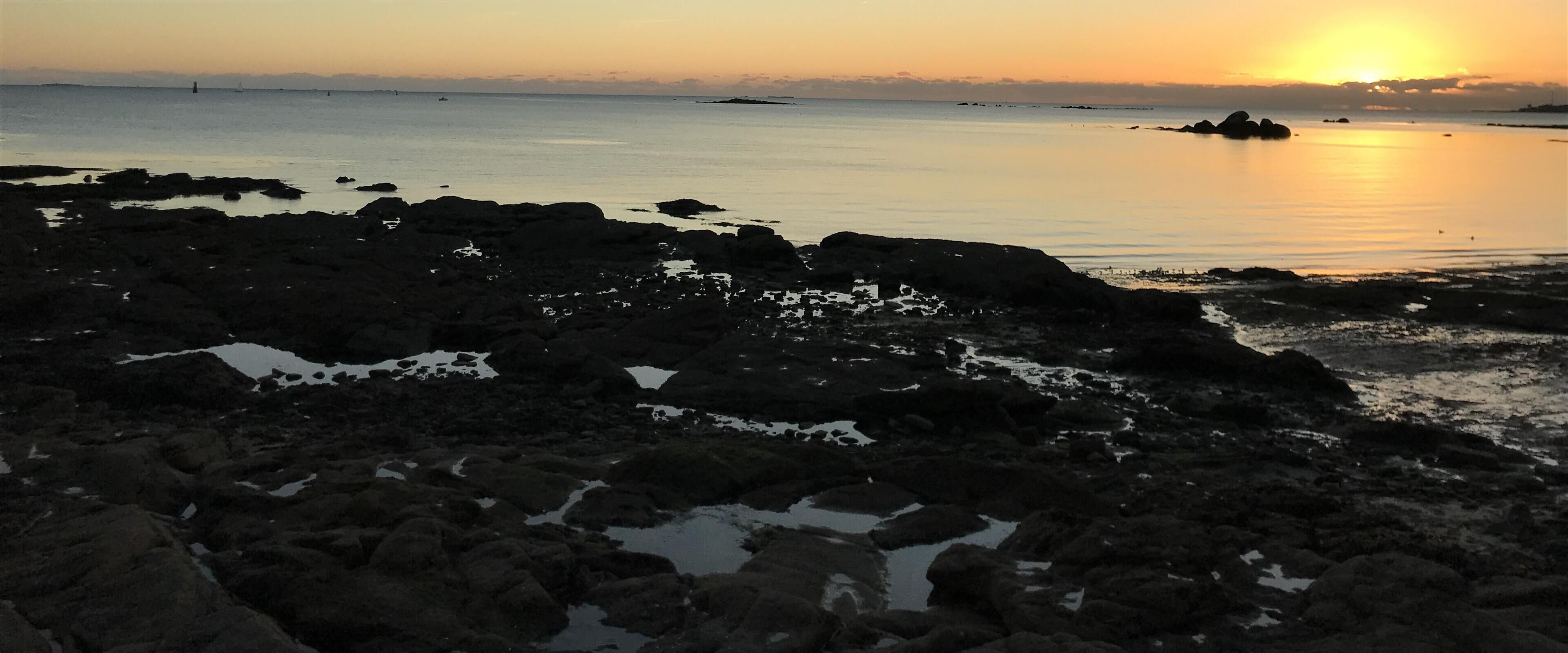 Une côte rocheuse bordée d’une eau calme, avec un coucher de soleil à l’horizon.