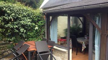 Een overdekt terras met een houten tafel en stoelen, een klein tafeltje met een tafelkleed met stippen, en uitzicht op de tuin door de glazen deuren.