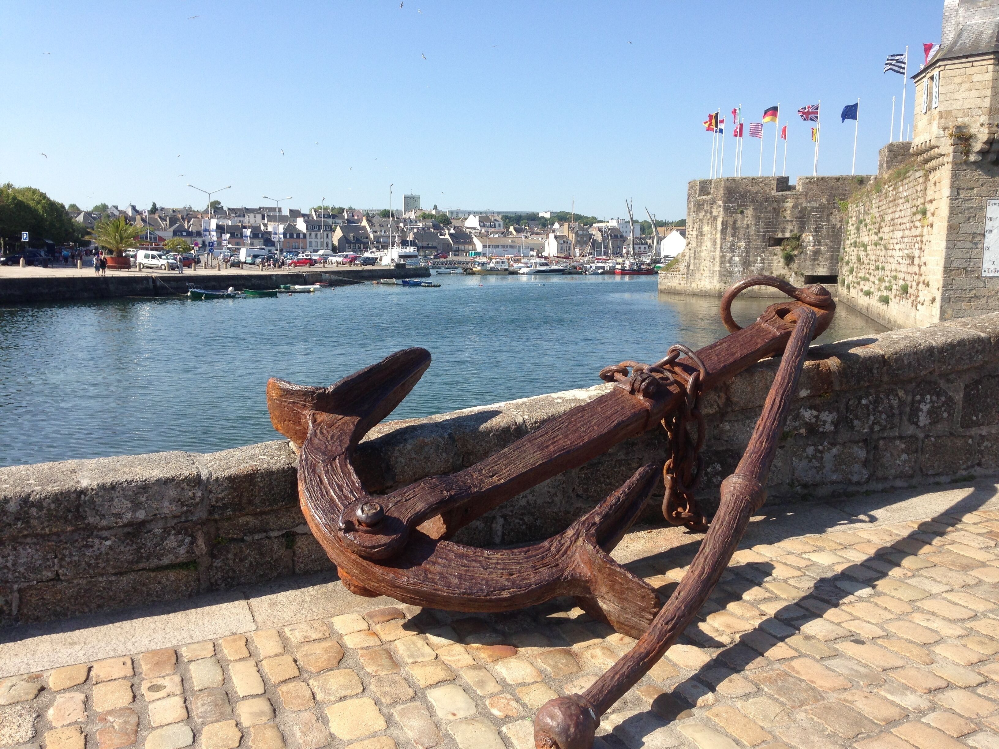 Une ancre fixée à un mur de pierre, au bord de l’eau, avec des bateaux et des drapeaux en arrière-plan.
