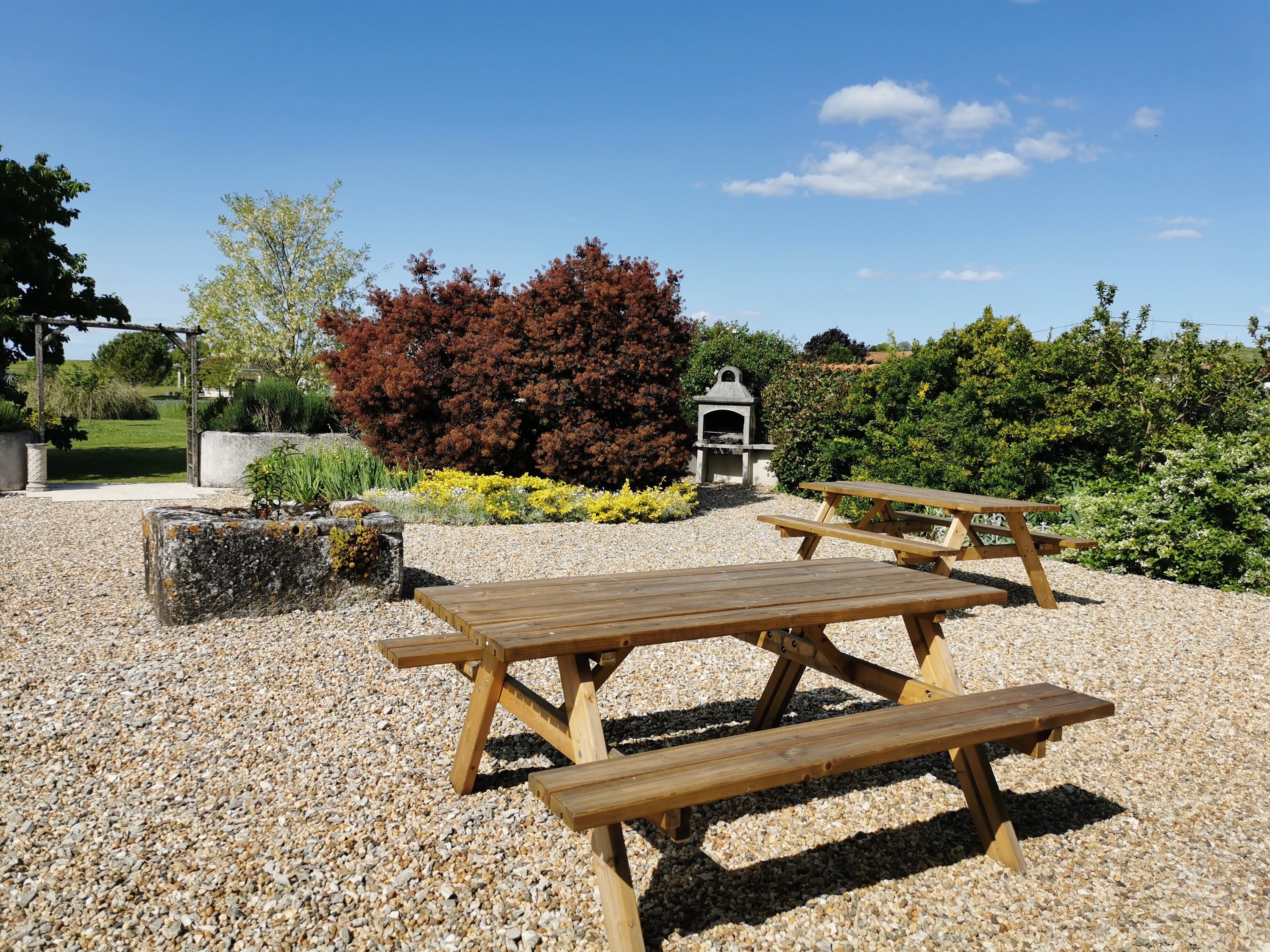 Ein Garten-Terrassenbereich mit Holztischen und -bänken, umgeben von Kies und verschiedenen Pflanzen.