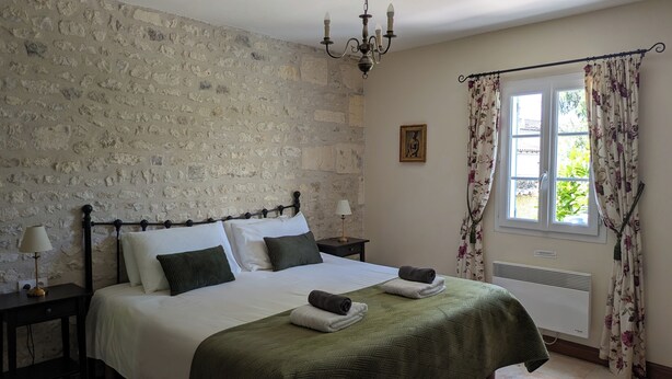 Een slaapkamer met een stenen muur, een bed met een groene sprei, een kroonluchter, een raam met bloemen gordijnen en een nachtkastje met een lamp.
