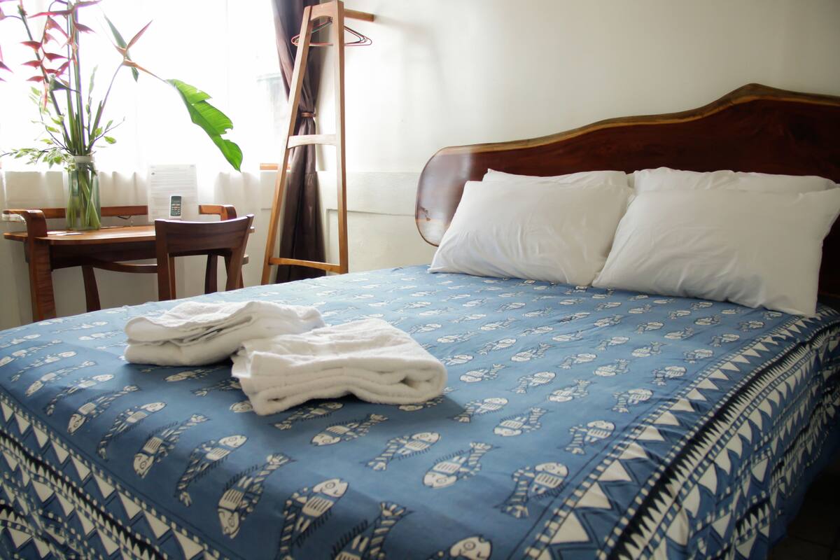Una cama bien hecha con una colcha azul estampada, un cabecero de madera y almohadas blancas. Se observa una silla de madera y una mesita con un florero y un teléfono.
