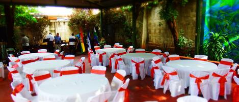 A covered outdoor event space with round tables and chairs, surrounded by greenery and a brick wall.