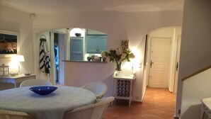A dining area with a round table, white chairs, a blue bowl, a small white cabinet with a vase of flowers, and a kitchen area in the background. - Villa 4 bedrooms, sleeps 300 meters from pampelone beach (Ramatuelle)