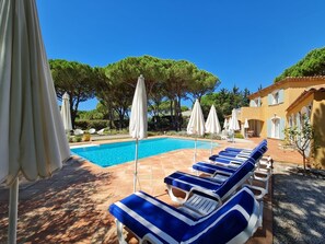 Un espace piscine agrémenté de chaises longues et de parasols, entouré d’arbres et bordé d’un bâtiment dont le toit est recouvert de tuiles.