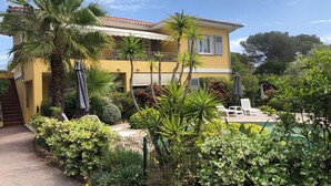 Um edifício amarelo com varanda, rodeado por vegetação exuberante e uma piscina.