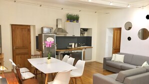 A modern living room with a grey sofa, white chairs, a wooden dining table, and a kitchen area with cabinets and a refrigerator. - Wonderful flat on the pear! (Nice)