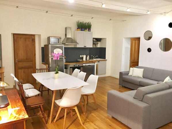 Ein modernes Wohnzimmer mit grauem Sofa, weißen Sesseln, einem Holzespeisetisch und einer Küchenzeile mit Schränken und einem Kühlschrank.