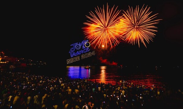 Evento nocturno al aire libre con fuegos artificiales, una multitud y un escenario con un letrero de neón.