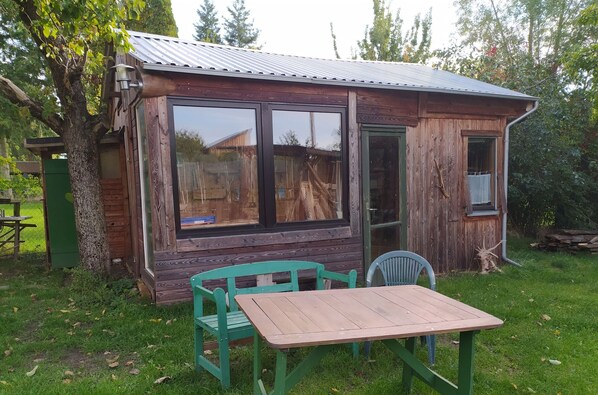 A rustic wooden cabin with a metal roof, large windows, and a small table with chairs in the yard.