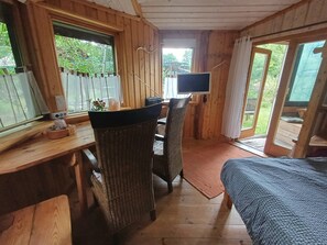 A wooden cabin interior with a bed, a desk, a chair, a television, and a view of the outdoors.