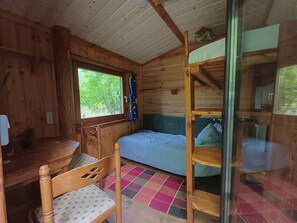 A wooden cabin interior with a bed, a chair, a table, and a window with a view of greenery.